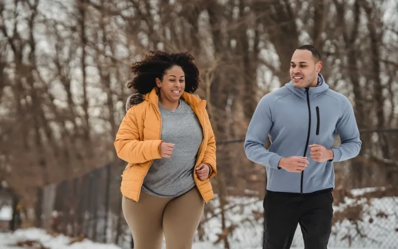Ein Mann und eine Frau joggen gemeinsam, es ist Winter. Sie trägt eine braune enganliegende Hose, ein graues Shirt und darüber eine orangenfarbene Dauenjacke, die geöffnet ist. Er trägt eine schwarze Hose und eine graue Fleecejacke mit Kapuze. Beide lächeln.