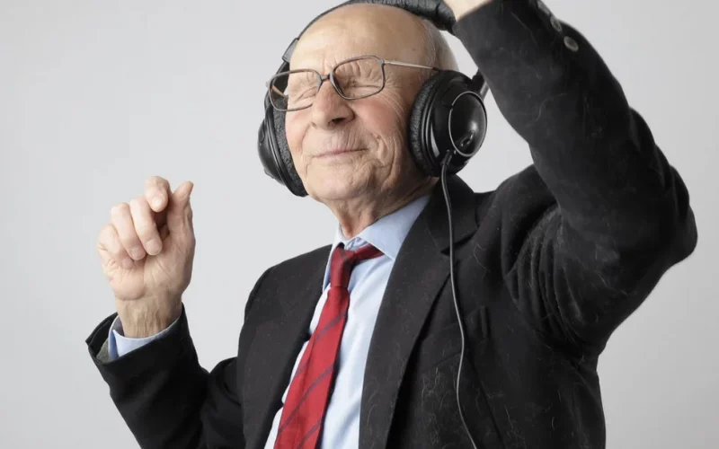 Ein älterer Herr mit Brille und wenigen Haaren, er trägt ein dunkles Sakko, hellblaues Hemd und eine rote Krawatte. Er hat schwarze Kopfhörer auf, die Augen geschlossen und hat die Hände in die Luft gestreckt. Er tanzt zur Musik.