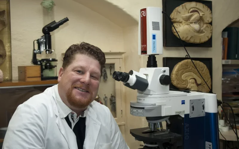 Prof. Dr. Dr. Jens Pahnke in seinem Labor vor einem Mikroskop. An der Wand im Hintergrund sind Darstsellungen des menschlichen Gehirns zu sehen.
