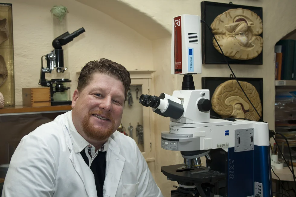 Prof. Dr. Dr. Jens Pahnke in seinem Labor vor einem Mikroskop. An der Wand im Hintergrund sind Darstsellungen des menschlichen Gehirns zu sehen.