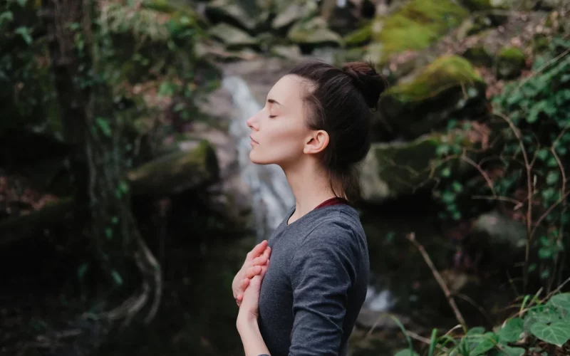Frau im Wald vor einem kleinen Wasserfall. Sie hat beide Hände auf Ihren Brustkorb gelegt und Augen geschlossen. Sie meditiert.