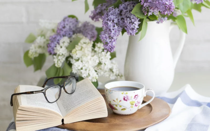 Buch, Brille und Teetasse auf einem kleinen Tisch mit einer Vase mit Flieder