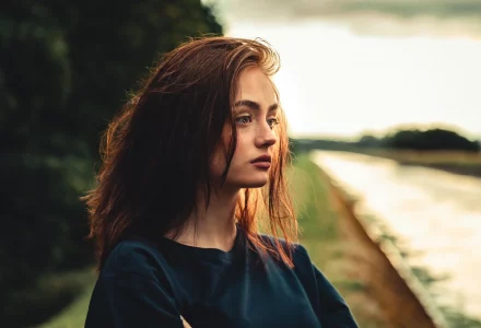 Ein junges Mädchen mit brünetten Haaren sieht melancholisch in die Umgebung. Die Umgebung, ein Fluss und ein Wald sind verschwommen.