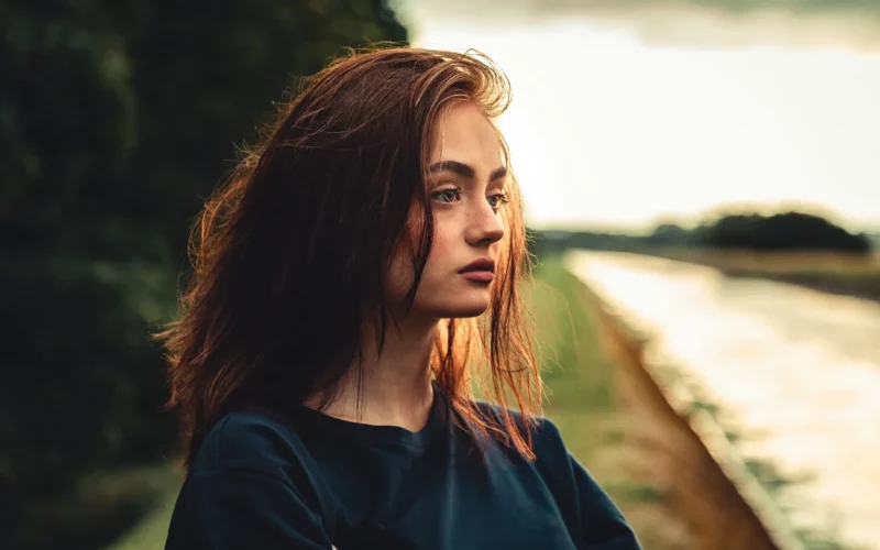 Ein junges Mädchen mit brünetten Haaren sieht melancholisch in die Umgebung. Die Umgebung, ein Fluss und ein Wald sind verschwommen.