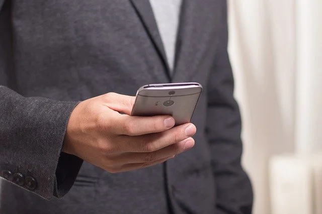 Ein Mann in dunkelgrauem Anzug hält ein Mobiltelefon in der Hand. Auf dem Bildausschnitt sind nur Oberkörper und Teil des rechten Armes und der Hand zu sehen. Der Oberkörper ist unscharf im Hintergrund. Hand mit Handy sind scharf im Vordergrund.